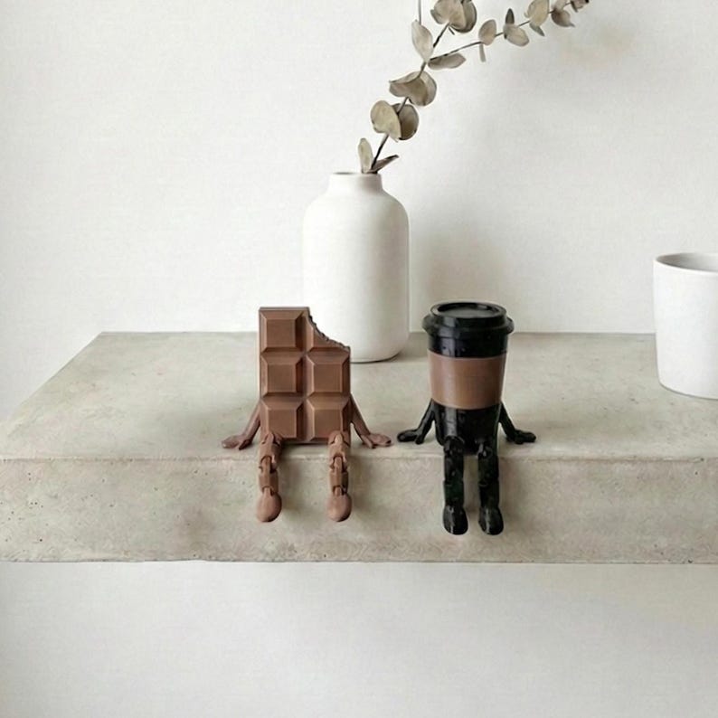 Pu&ograve; includere: Un'eccentrica serie di figurine di cioccolata e tazza di caff&egrave;. La barretta di cioccolato &egrave; marrone con un morso mancante, e la tazza di caff&egrave; &egrave; nera e marrone. Entrambi hanno braccia e gambe, dando loro un aspetto giocoso. Un vaso bianco con un rametto di verde &egrave; sullo sfondo.