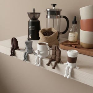 May include: A collection of whimsical coffee-themed figurines, including a coffee bean, a stovetop espresso maker, a coffee cup, a chocolate bar, and a to-go coffee cup, each with legs. A coffee grinder, French press, and coffee beans in a burlap sack are in the background.