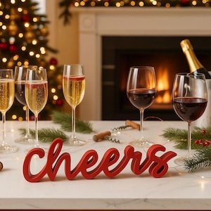 Peut inclure: Une scène festive avec des verres de vin remplis de vin rouge et de vin mousseux. Un signe rouge "cheers" est placé devant les verres. Un sapin de Noël et une cheminée sont en arrière-plan, créant une ambiance chaleureuse et festive.
