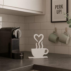Könnte beinhalten: Eine schwarze Espressomaschine steht auf einer Arbeitsplatte neben einer weißen dekorativen Kaffeetasse mit herzförmigem Dampfdesign. Zwei grüne Tassen hängen an der Wand, und ein gerahmtes Schild zeigt "PERK UP!"