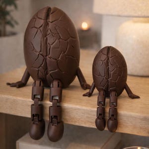 May include: Two brown, egg-shaped sculptures with articulated legs and arms sit on a wooden surface. The larger sculpture is approximately 10 inches tall, while the smaller one is about 6 inches tall. Both have a cracked texture.