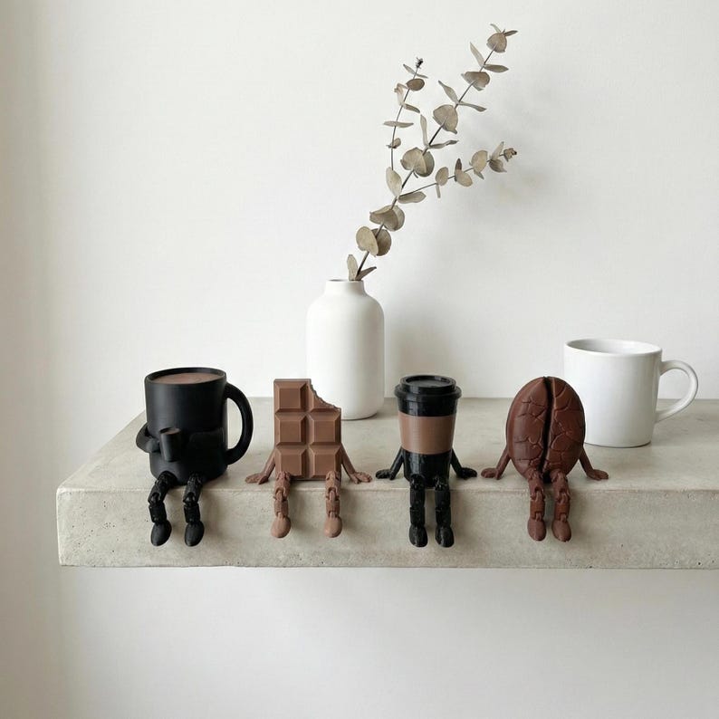 Pu&ograve; includere: Una tazza da caff&egrave; nera, una barretta di cioccolato con un morso, una tazza da caff&egrave; e un chicco di caff&egrave;, ciascuno con gambe e braccia, sono disposti su una mensola. Sono presenti anche un vaso bianco con un rametto verde e una tazza bianca.