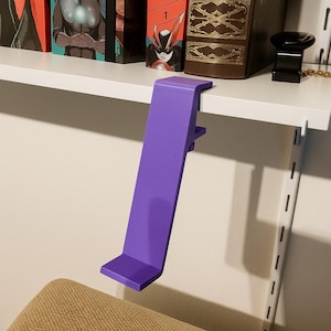 May include: A vibrant purple shelf clip, designed to attach to a shelf. The clip is made of a solid material and has a rectangular shape with a right-angle bend at the top and bottom. It is designed to hold items on a shelf.
