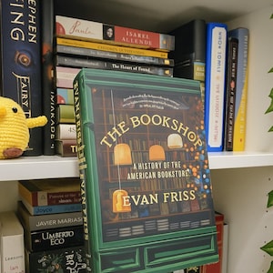 May include: A hardcover book titled "The Bookshop: A History of the American Bookstore" by Evan Friss. The cover is green with a bookstore illustration and the title in gold. The book is on a shelf with other books.