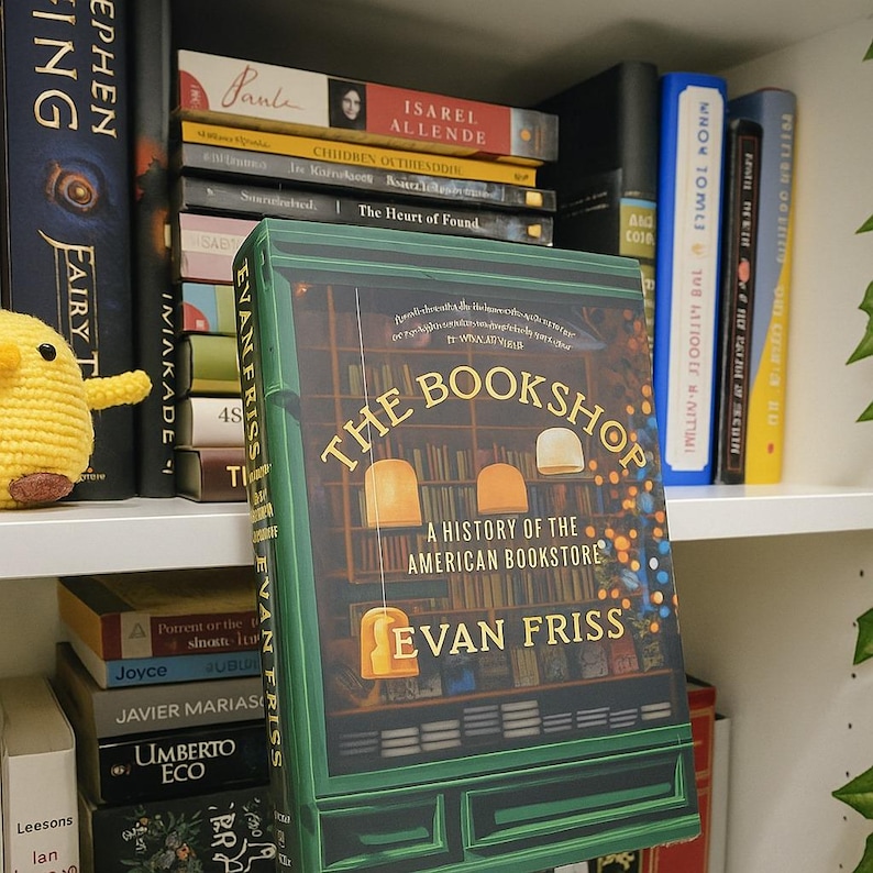 May include: A hardcover book titled "The Bookshop: A History of the American Bookstore" by Evan Friss. The cover is green with a bookstore illustration and the title in gold. The book is on a shelf with other books.