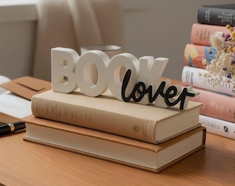 May include: A decorative sign spelling "BOOK lover" in white and black, placed on two stacked books with beige covers. The scene is set on a wooden table, with other books visible in the background.