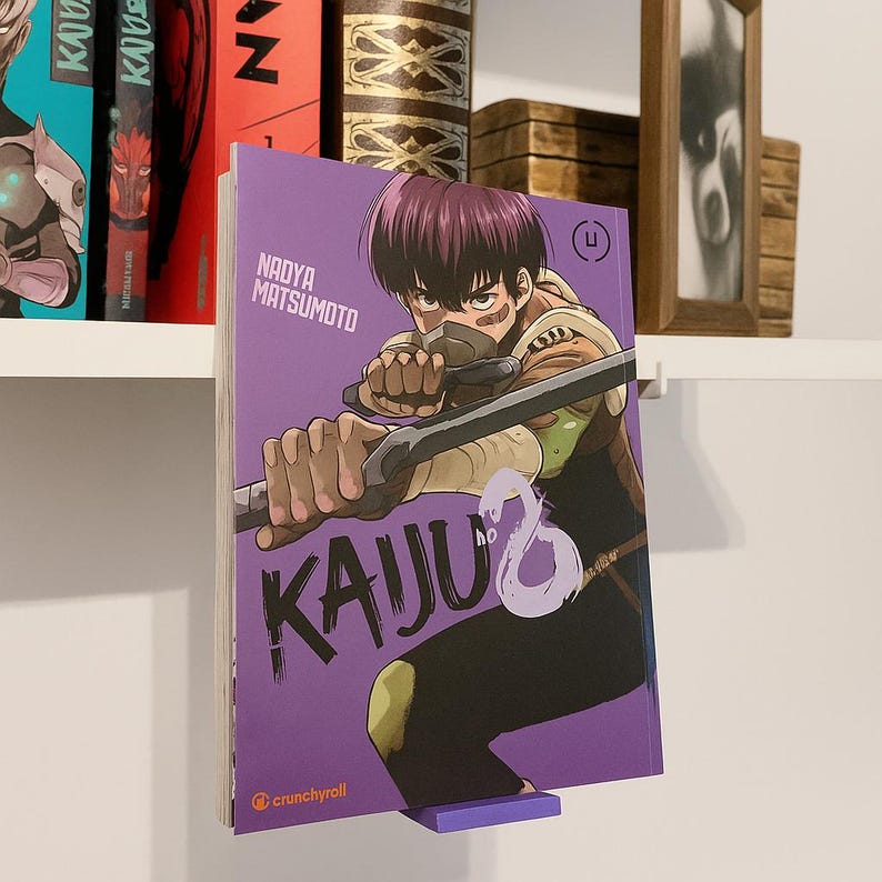 May include: A purple manga volume titled "KAIJU" by Nadya Matsumoto, featuring a character with a sword. The book is displayed on a purple stand. Other manga volumes are visible on a shelf in the background.