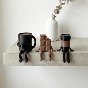 Pu&ograve; includere: Tre figurine stravaganti sedute su una mensola. Una tazza da caff&egrave; nera, una barretta di cioccolato con un morso e una tazza da caff&egrave; con un coperchio. Ognuna ha braccia e gambe, conferendo loro un aspetto giocoso e antropomorfo. Un vaso bianco con un rametto di verde &egrave; sullo sfondo.