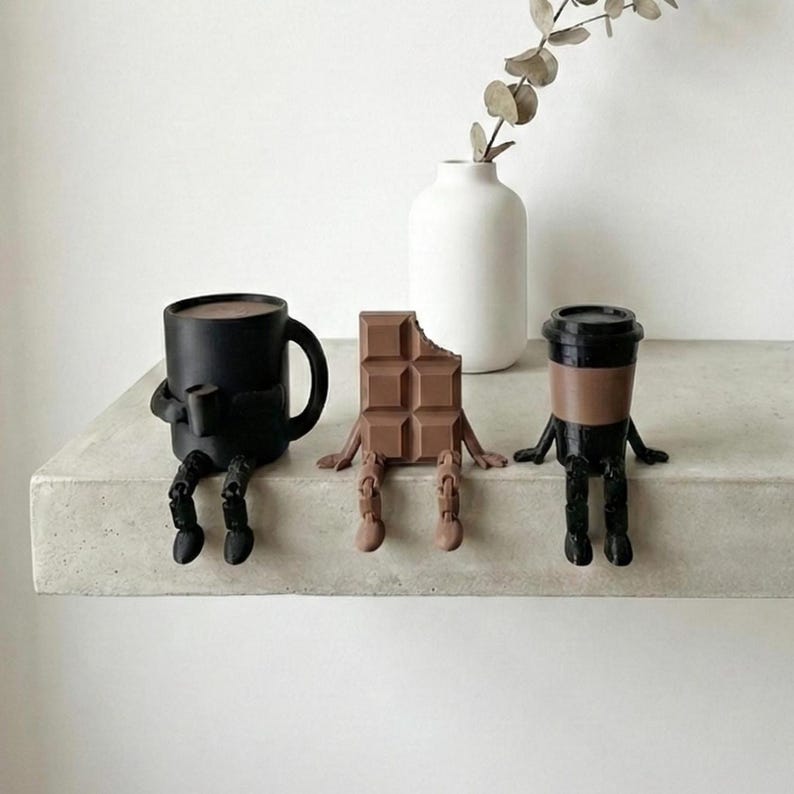 Pu&ograve; includere: Tre figurine stravaganti sedute su una mensola. Una tazza da caff&egrave; nera, una barretta di cioccolato con un morso e una tazza da caff&egrave; con un coperchio. Ognuna ha braccia e gambe, conferendo loro un aspetto giocoso e antropomorfo. Un vaso bianco con un rametto di verde &egrave; sullo sfondo.