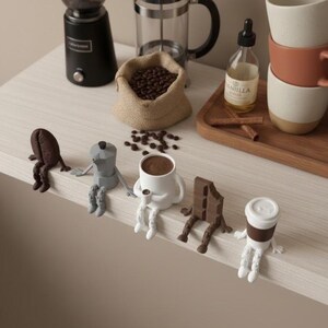 May include: A coffee-themed still life featuring a black coffee grinder, a French press, a burlap sack of coffee beans, and various coffee-related figurines. A bottle of vanilla extract, cinnamon sticks, and stacked mugs are also present.