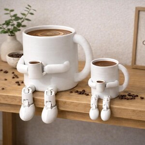 May include: Two white ceramic coffee mugs with a whimsical design. The larger mug holds a full cup of coffee and a smaller mug. Both mugs have arms and legs, giving them a playful, anthropomorphic appearance. Coffee beans are scattered around.