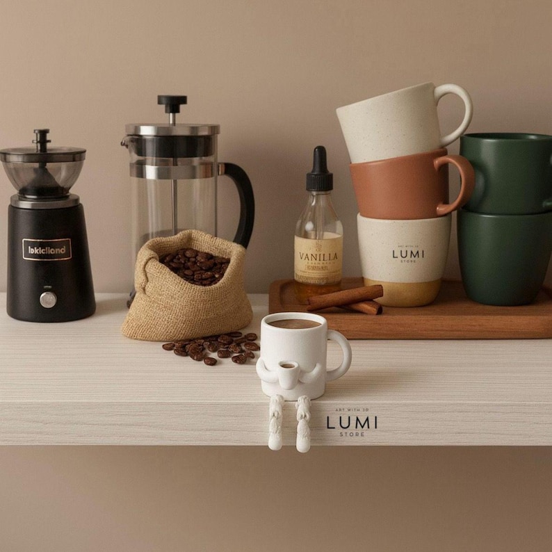 May include: A coffee-themed still life featuring a black coffee grinder, a French press, a burlap sack of coffee beans, and a bottle of vanilla extract. Several stacked mugs in various colors and a whimsical mug with legs are also present. The Lumi Store logo is visible.