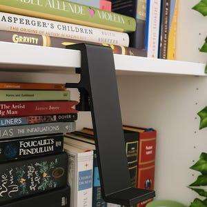 May include: A black metal shelf clamp is attached to a white shelf, with a stack of books visible. The clamp has a right-angle design with a screw mechanism for securing it to the shelf. The books have various colors and titles.
