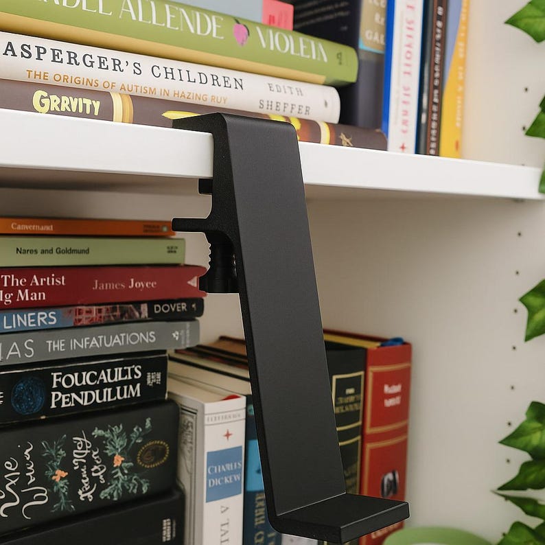 May include: A black metal shelf clamp is attached to a white shelf, with a stack of books visible. The clamp has a right-angle design with a screw mechanism for securing it to the shelf. The books have various colors and titles.