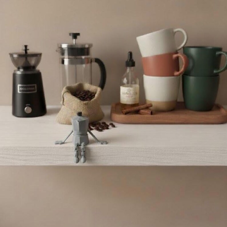 Pu&ograve; includere: Un'attrezzatura per il caff&egrave; con un macinacaff&egrave; nero, una French press, un sacco di chicchi di caff&egrave; e una bottiglietta. Tazze impilate in bianco, terracotta e verde su un vassoio di legno. Una statuina grigia di caffettiera espresso in primo piano.