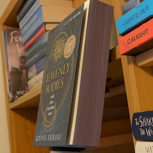 May include: A hardcover book titled "Heavenly Bodies" with a dark blue cover and gold accents. The book is displayed on a wooden shelf with other books. The spine is a deep purple color. The author's name, Kinaas Errim, is visible.