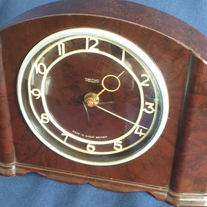 Może przedstawiać: Zegar w stylu vintage w kolorze brązowym z zakrzywioną górą i okrągłą tarczą. Tarcza zegara ma białe cyfry i wskazówki, z widoczną nazwą marki "Smiths". Zegar wyprodukowano w Wielkiej Brytanii.