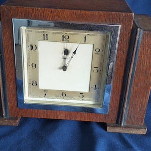 Op de afbeelding: Een vintage houten klok met een rechthoekige wijzerplaat en een zilverkleurig frame. De wijzerplaat is gebroken wit met zwarte cijfers en wijzers. De klok is omhuld in een donkerbruin houten frame met een trapvormig ontwerp.