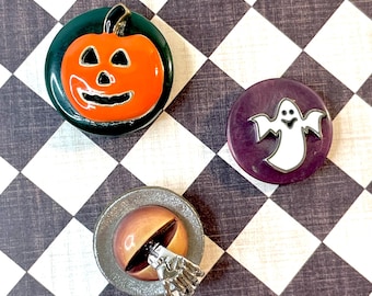 May include: Three Halloween-themed pins. One pin features an orange jack-o'-lantern on a green background. Another pin has a white ghost on a purple background. The third pin has a brown eyeball with a silver hand.