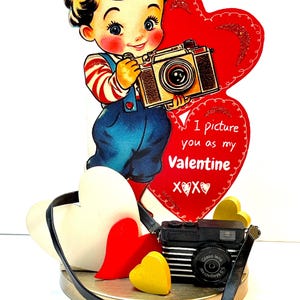 May include: A vintage Valentine's Day card featuring a smiling boy holding a camera. The boy is wearing blue overalls and a red and white striped shirt. The card includes a red heart with the text "I picture you as my Valentine" and a small black camera with a strap.