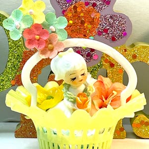 May include: A vintage figurine of a girl in a yellow basket filled with flowers. The basket has a white handle and is surrounded by colorful floral decorations. The figurine is white with a serene expression.