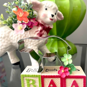 May include: A metal lamb-shaped cookie cutter rests on wooden blocks spelling "BAA". A ceramic lamb planter with flowers is in the background. The blocks are green and red with the letters in matching colors. Small paper flowers are attached to the blocks.