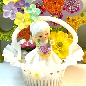 May include: A vintage figurine of a girl in a white dress holding a basket of flowers. The figurine is set inside a white cupcake liner, surrounded by colorful paper flowers and glittery floral decorations. The overall theme is whimsical and decorative.