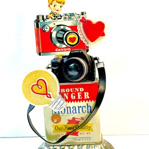 May include: A whimsical assemblage featuring a red and silver "CANDID" camera, a black camera, and a vintage "Monarch" food container. Adorned with a heart-shaped decoration and a lightbulb with a heart. The base is a decorative silver dish.