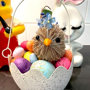 May include: An Easter-themed decorative basket shaped like a cracked egg, filled with colorful speckled eggs and a straw chick with blue floral accents. A white handle arches over the basket. A yellow figurine and a white bunny figurine are in the background.