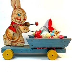 May include: Vintage Easter toy featuring a rabbit pulling a blue wagon filled with Easter eggs and other toys. The rabbit is made of cardboard and has a red vest. The wagon has yellow wheels.
