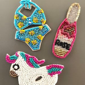 May include: Three beaded accessories: a turquoise bikini with yellow flower accents, a pink and gold champagne bottle, and a white unicorn with pink and turquoise mane and tail. The items are embellished with beads.