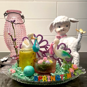 May include: A springtime centerpiece featuring a lamb figurine, stork figurines, and a glass dish with decorative elements. The arrangement includes a teal speckled egg, colorful flowers, and the word "SPRINGTIME" in multi-colored letters.