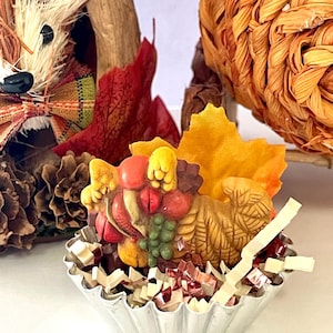 May include: A small cornucopia figurine filled with miniature red, yellow, and green fruit, nestled in a silver fluted metal tart tin with shredded paper filler and a single orange fabric maple leaf extending from the back.