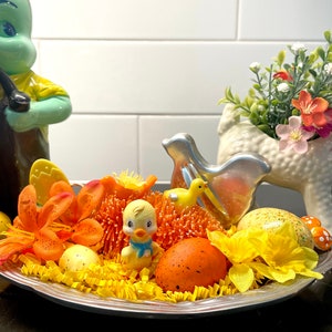 May include: A decorative Easter display featuring a silver tray filled with yellow paper grass, orange flowers, and speckled eggs. A yellow chick figurine with a blue bow and a small yellow duck are also present. A green bunny figurine is in the background.