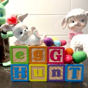 May include: A whimsical Easter-themed display featuring a white ceramic bunny with a flower crown, colorful eggs, and wooden blocks spelling "EGG HUNT." A lamb figurine and a green bunny figurine are also present.