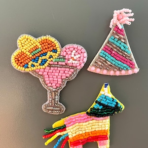 May include: Three beaded patches: a pink margarita glass, a party hat with pink and blue stripes, and a colorful piñata. The patches are made with small, colorful beads and silver metallic thread.