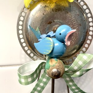 May include: A whimsical, handmade bird ornament. The blue bird has a pink beak and is perched on a wooden bead. A green and white gingham ribbon and a yellow flower add decorative touches. The ornament is attached to a metal plate.