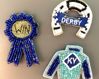 May include: Three beaded horse racing themed pins. A blue ribbon with "WIN" in gold, a horseshoe with "DERBY" on a blue banner, and a turquoise and blue jockey shirt with "KY" on the chest.