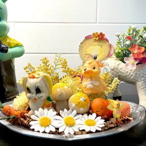 May include: A silver tray holds Easter-themed decorations. A white bunny figurine with a carrot, yellow duckling, and mushroom figurines are surrounded by daisies, eggs, and yellow floral accents. The arrangement is festive and colorful.