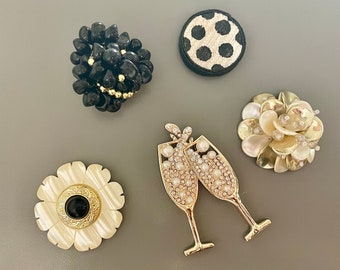 May include: An assortment of decorative brooches. Includes a black stone cluster, a black and white polka dot circle, a flower with a black center, a champagne glass design, and a flower with pearl accents.