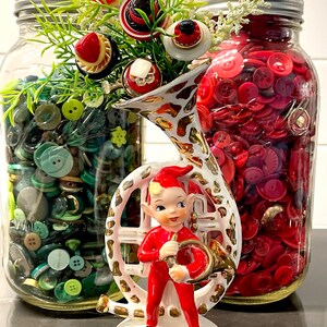 May include: A whimsical arrangement featuring a vintage ceramic elf figurine in a red suit holding a golden horn, flanked by two glass jars filled with colorful buttons. One jar holds green buttons, the other red.