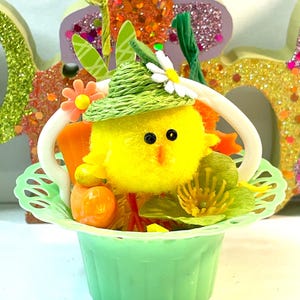 May include: A decorative Easter basket featuring a fluffy yellow chick wearing a green hat with a white daisy. The basket is green with a white handle and filled with orange carrots and green foliage.