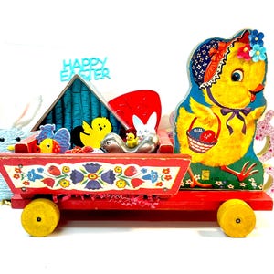 May include: A vintage red wooden wagon with yellow wheels, filled with Easter-themed decorations. The wagon features floral designs and carries a yellow chick, a silver bunny, and other festive items. A large cardboard chick stands at the end.
