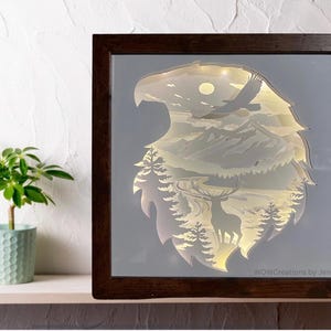 Pode incluir: Uma moldura de madeira com fundo branco e uma silhueta de uma águia com um veado e montanhas dentro. A cabeça da águia está voltada para o lado esquerdo da moldura. O texto "WOWCreations by Jen" está no canto inferior direito da moldura.