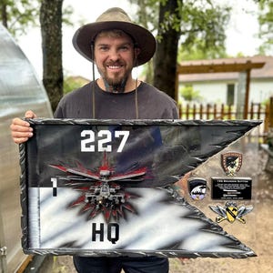 May include: A black and white flag with the numbers "227" and "HQ" displayed prominently. The flag features a detailed graphic of a helicopter and several military patches. The flag is framed in a black, textured border.