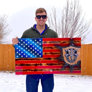 May include: A wooden American flag with a burnt effect and a special forces insignia in the lower right corner. The flag is held by a person wearing sunglasses and a green jacket.