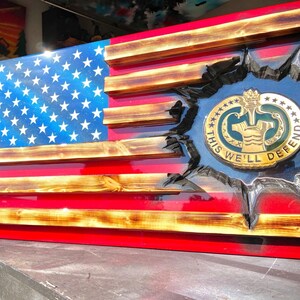May include: A wooden American flag with a distressed finish. The flag is mounted on a red background and has a gold-colored military emblem in the center. The emblem has the words "This We'll Defend" around the edge.