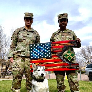 May include: Two people in military uniform stand in front of a wooden American flag with a green patch. A white and grey husky dog sits in front of them.