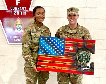 Pode incluir: Duas pessoas em uniforme militar estão segurando uma bandeira americana de madeira com um visual desgastado. A bandeira tem um efeito queimado e um selo do Exército dos Estados Unidos no canto inferior direito. A bandeira está montada em uma placa de madeira.