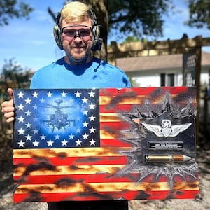 May include: Wooden American flag with a blue star field and red and white stripes. The flag features a helicopter image, military wings, and a bullet. The flag is approximately 24 inches wide and 12 inches tall.